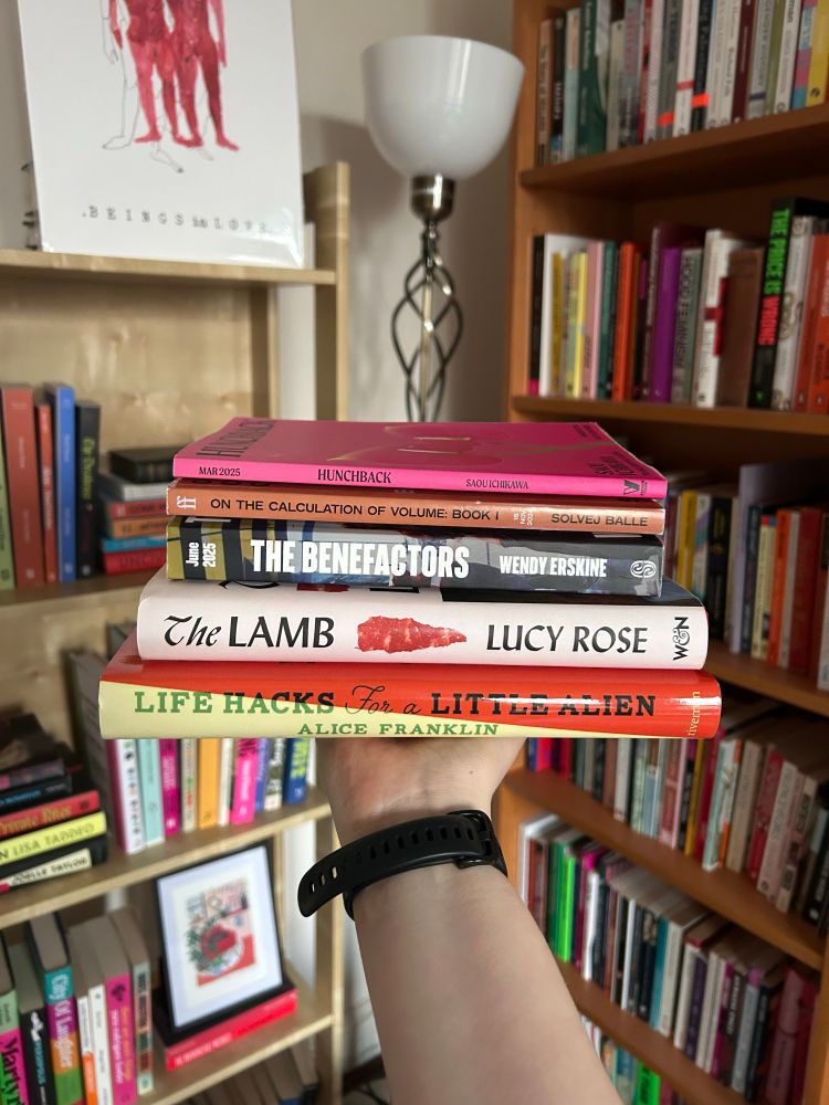 my hand is holding Alice Franklin’s Life Hacks for a Little Alien, Lucy Rose’s The Lamb, Wendy Erskine’s The Benefactors, Solvej Balle’s On the Calculation of Volume, Saou Ichikawa’s Hunchback. the background is bookshelves