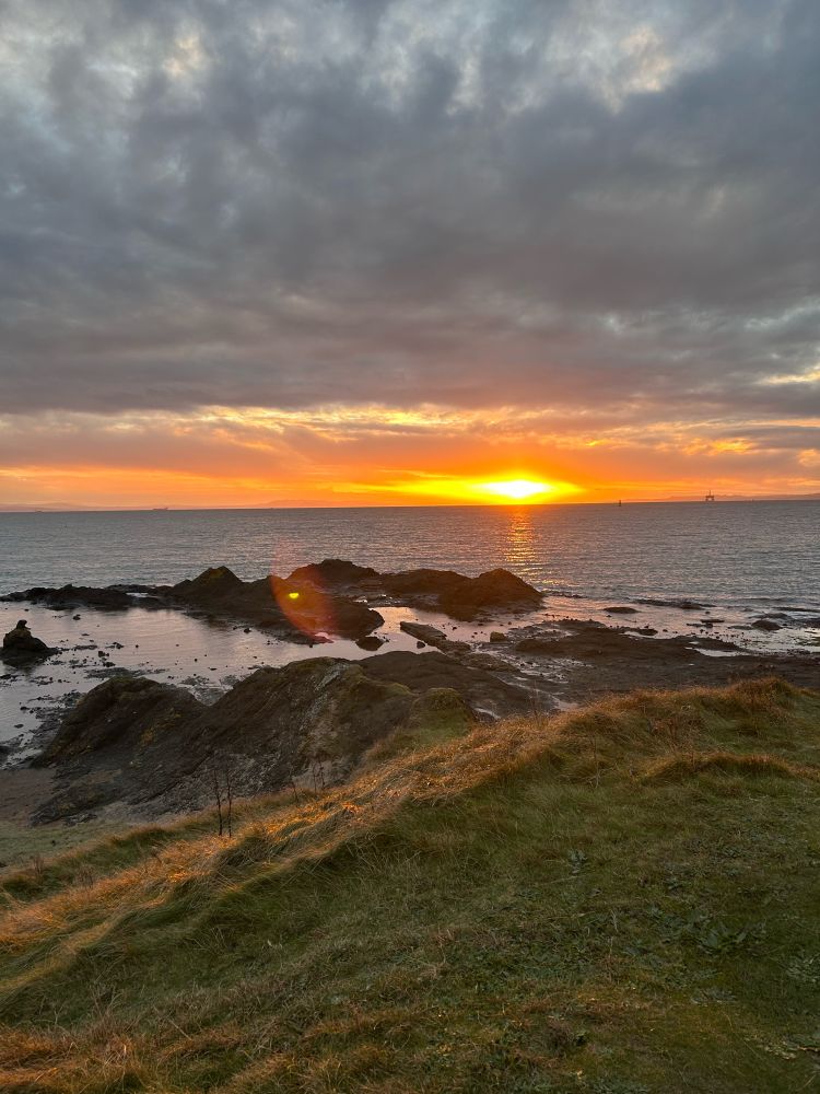 The sun setting over the water. Rocks and a grassy bank are in the foreground. The sky is cloudy 
