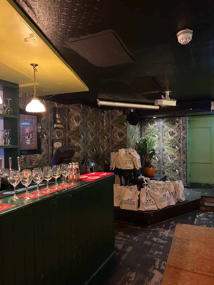 a photo of a room in a pub. there are many glasses on the bar and, in the background, a large stack of Hodder & Stoughton branded totes filled with books