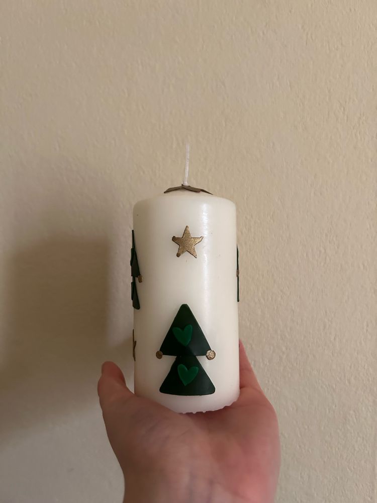 the user’s hand is holding a thick white candle decorated with gold stars and christmas trees with gold baubles 