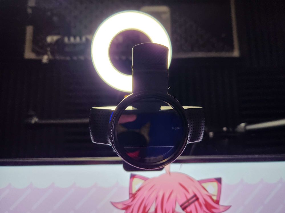 The image shows a webcam with a circular polarizer (CPL) filter attached to the lens. The webcam is positioned in front of a bright ring light, which is illuminating the scene from behind, creating a glowing halo effect around the filter. In the background, the room appears to be set up with acoustic panels for soundproofing. The lower part of the image reveals part of a computer screen with an anime or virtual character featuring pink hair and cat ears.