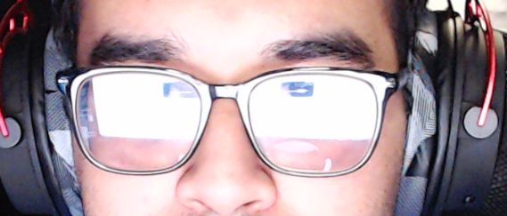 The image shows a close-up of a person's face wearing glasses and over-ear headphones. The reflection of a computer screen is visible in the lenses of the glasses. The lighting emphasizes the reflection, while the person's facial features, such as their eyebrows and part of their forehead, are in focus.