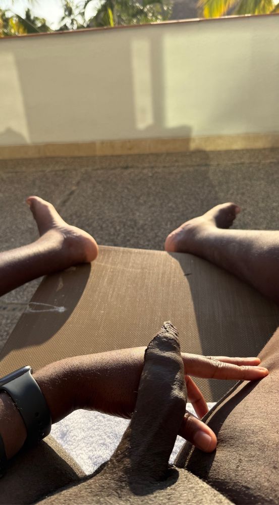 Tug sits on a patio chair during golden hour with his semi erect penis resting on his left hand with his foreskin providing full coverage and dangling off the tip. Both of his feet dangle of the chair in the background.