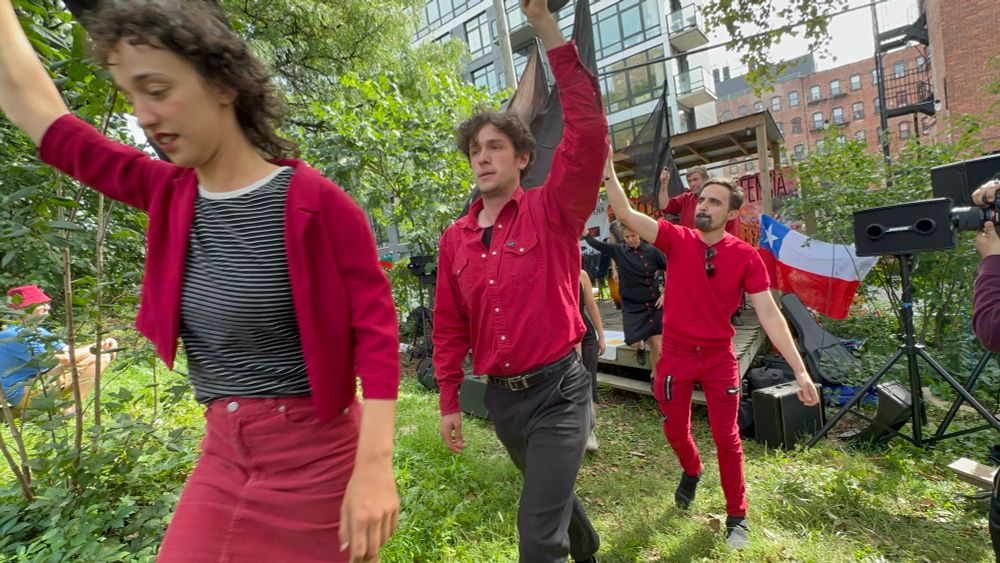 Green grass and leaves below and on the sides and a large brick apartment in the background. A young woman, followed by two young men, march away from the stage and toward the massed crowd, each one with a hand raised and wearing red - the woman with a crimson cardigan and skirt, in the middle a young man with a long sleeved scarlet double breast pocket shirt with white buttons, and behind him a man with a goatee, red t-shirt and jeans, shades hanging from his collar, just below his chin. They are performing in an event in the Bronx to mark the anniversary of Salvador Allende's martyrdom in September. 