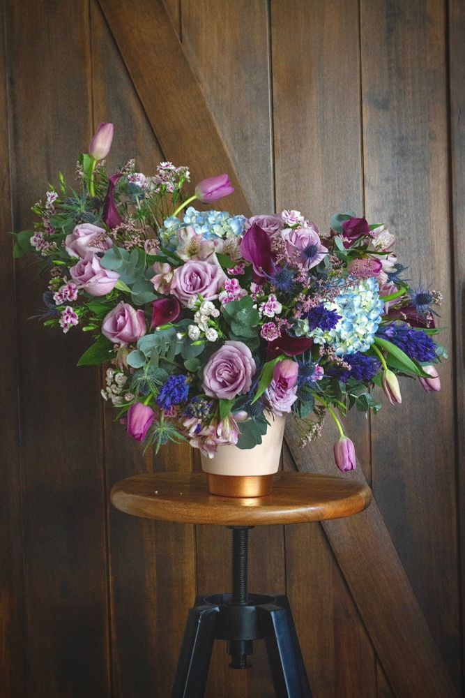 A large floral arrangement in tones of purple and blue sits on top of an industrial stool with a dark brown wooden barn door behind it.