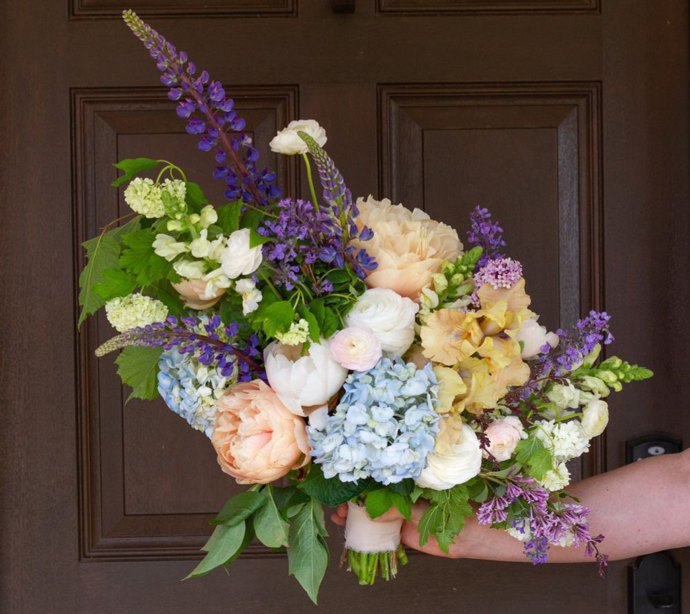 A hand holds up a pastel toned bridal bouquey in tons of peach, cream, pale blue, and purple. There is a dark brown door behind it.