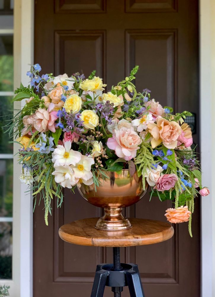 A large floral arrangement in pastel tones sits in a copper pedestal dish on top of an industrial looking stool.  