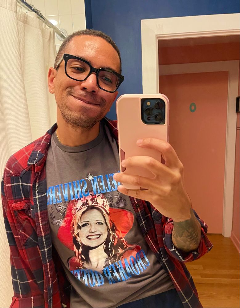 A man (me!) takes a mirror selfie with a pink phone. He’s wearing a red and blue flannel over a Helen Shivers Croaker Queen shirt. It’s the same design used in I Know What You Did Last Summer movie. He’s happy. 