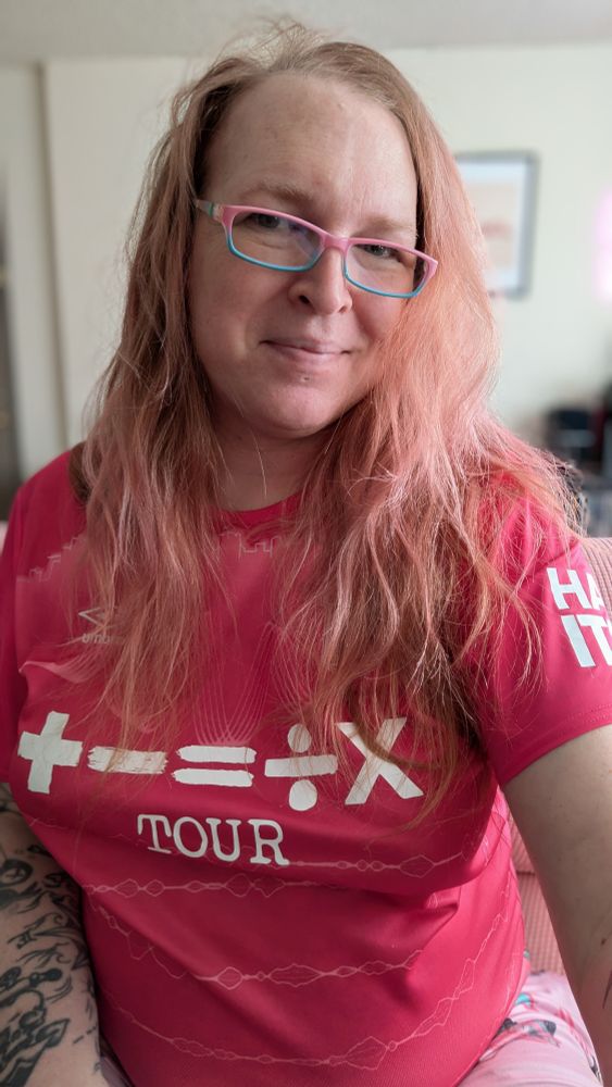 A middle-aged white woman with long blonde hair wearing a pink Ipswich Town FC kit and pink and blue reading glasses 