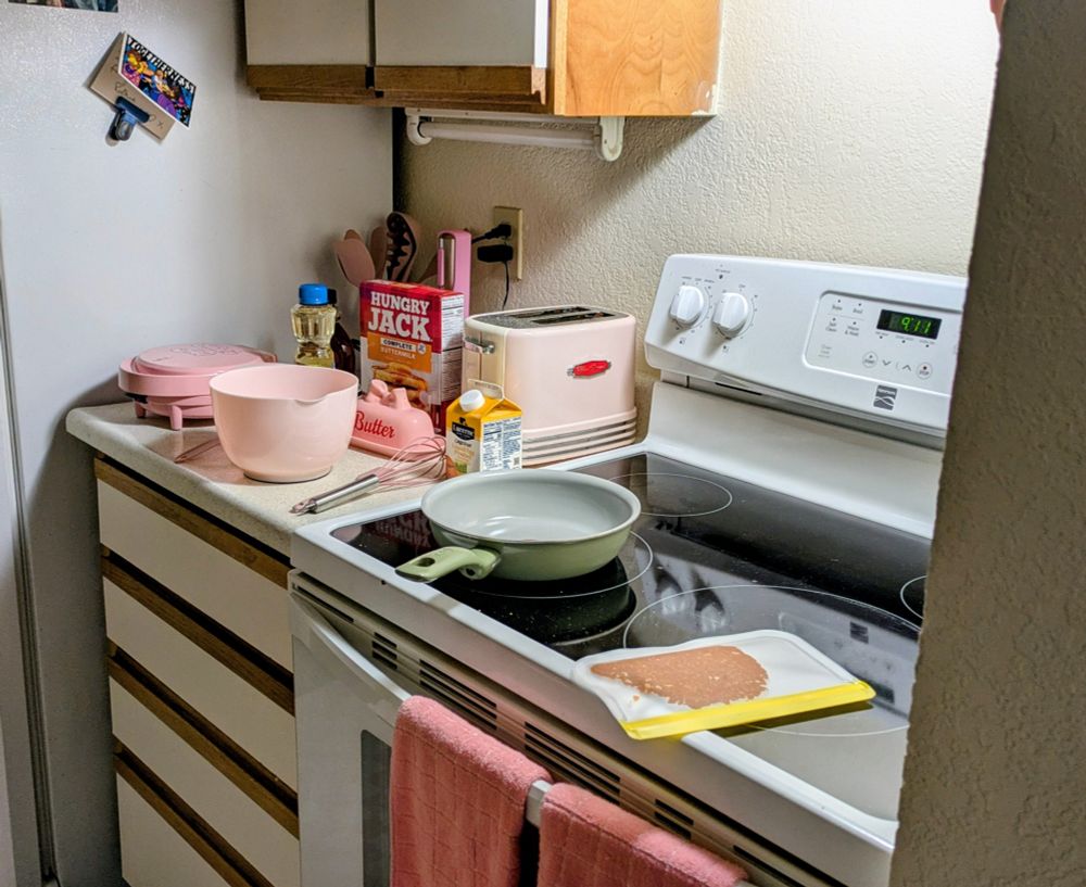 A white stove and beige kitchen counter. On the stove is a green T-Fal pan and a ziplock bag of soyrizo. On the oven for handle are two pink hand towels. 

On the counter is a Paris Hilton brand waffle maker and assorted waffle making supplies. There's also a pink, retro looking toaster but that's not relevant to what's happening here.