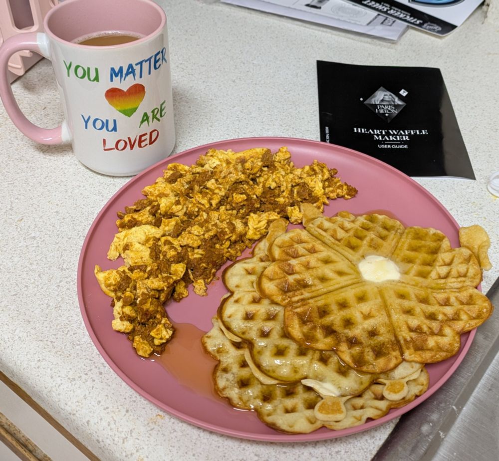 A pink plate with 3 waffles with heart-shaped segments and scrambled soyrizo eggs. A cup of coffee is to the left in a pink and white mug with the words "You Matter, You Are Loved" and a rainbow heart on it.