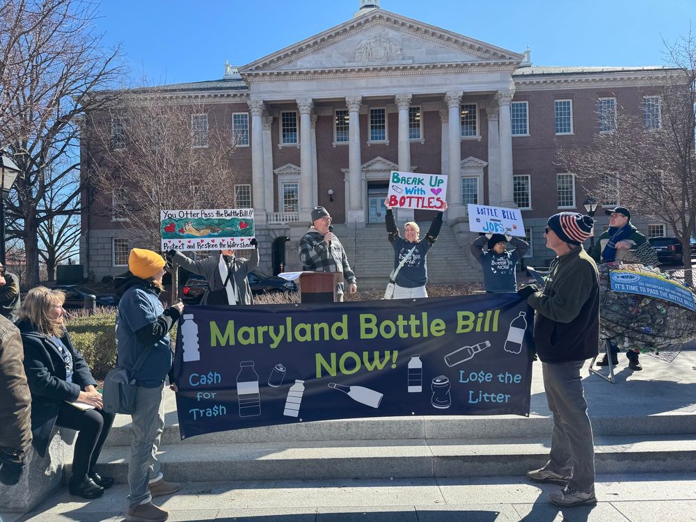 People wearing shirts that say bottle bill now stand in front of MD state house and hold up handmade signs behind a banner that says bottle bill now. Handmade signs say you otter pass the bottle bill, break up with bottles, and bottle bill jobs.