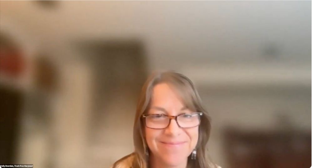 Screen shot of a woman wearing glasses and bottle earrings with a blurred background in a zoom meeting.