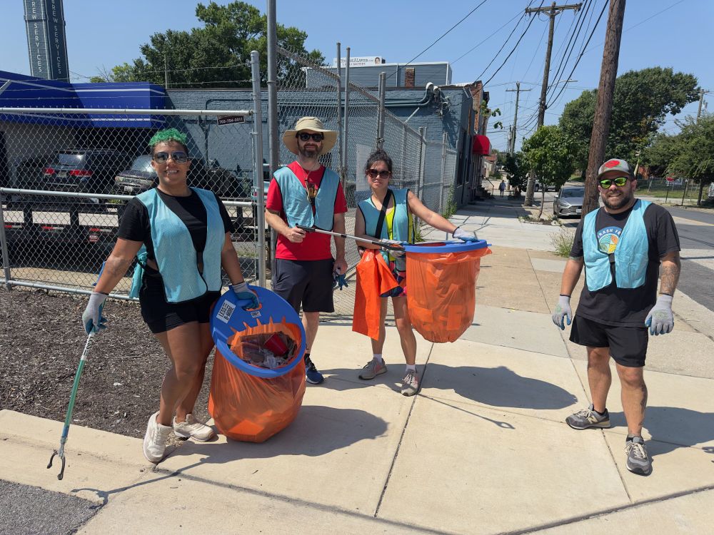 Picture shows 4 adults on an urban sidewalk holding trash grabber tools and carrying orange trash bags and wearing blue safety vests under a hot sun.
