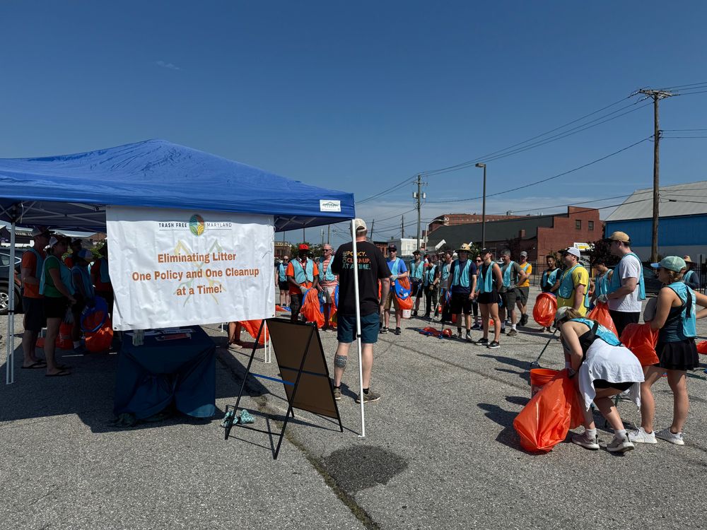 Photo shows group of 70 people wearing blue safety vests and holding orange trash bags and listening to a man with a megaphone in front of a tent and banner that says, "Trash Free Maryland - Eliminating Litter One Policy and One Cleanup at a Time."