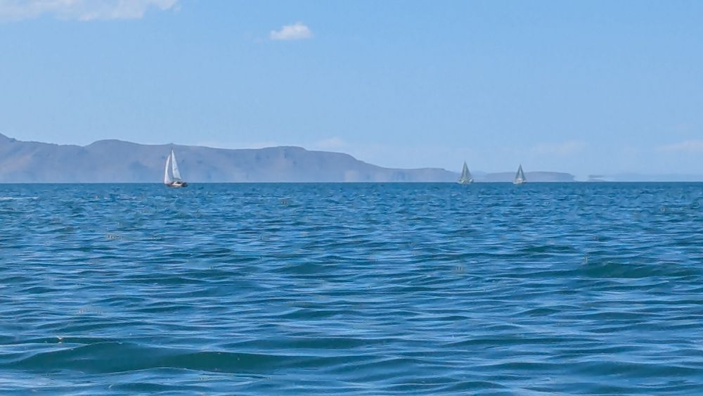 Three sailboats in the distance under sail. Mountains stand in the far distance peaking above the water line.
