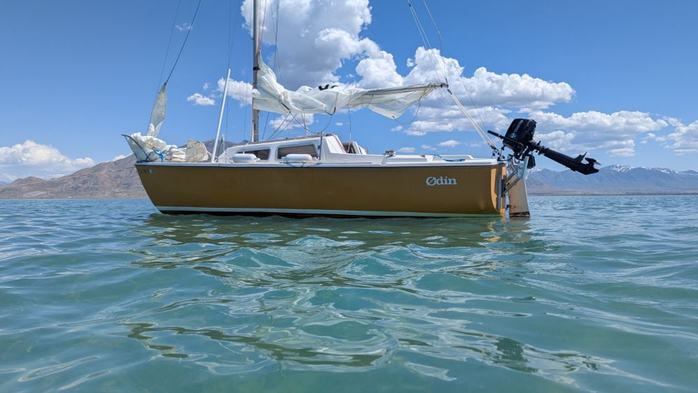 An orange Catalina 22 Sailboat sits at anchor under a brihg sky. Mountains rise from the background with large billowing clouds dotting the sky. 