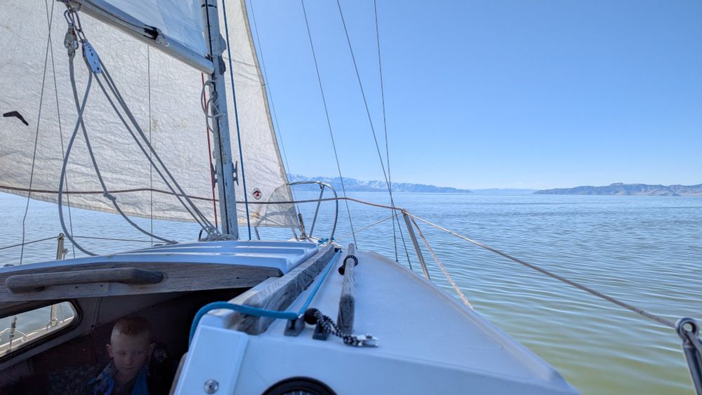 A sailboat sails over a large lake. Snow capped mountains line the horizon.