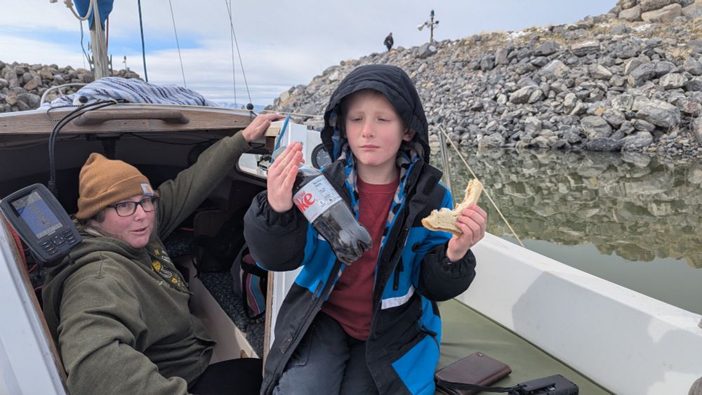 A view on a sailboat with mounds of rocks making up the mouth of a marina. A young boy eats his sandwich as he hands back a bottle of diet Coke. His mother sits below in the cabin to hand things out.