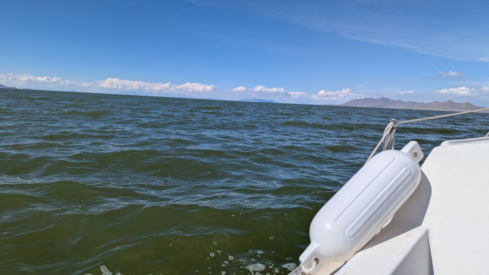 A view from a sailboat in the water. The fender is slipped onto the boat. In the distance, rain can be seen coming down along the shore, yet it is sunny on the ship. 