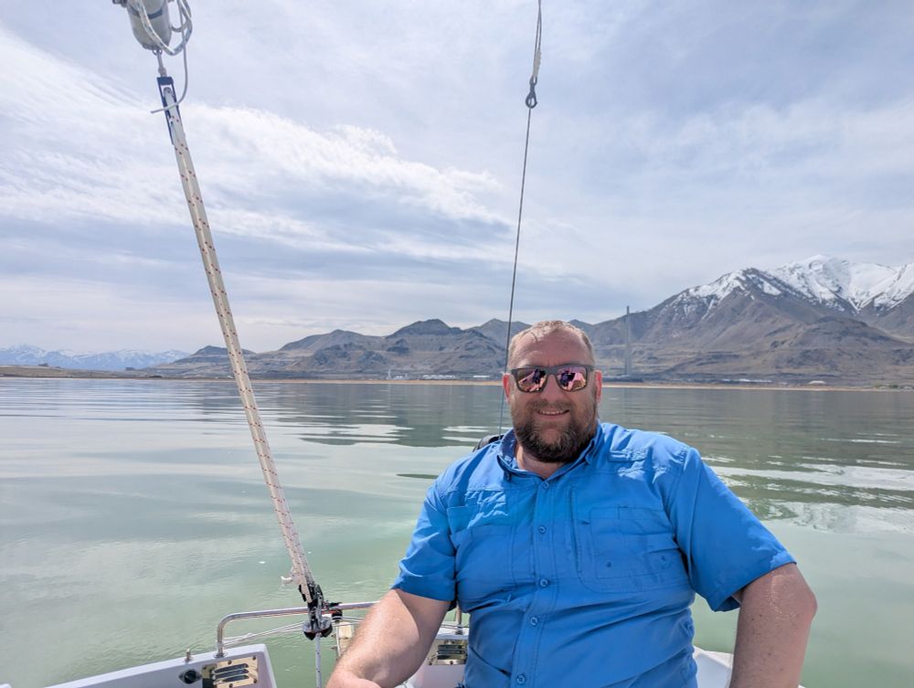 A middle age sexy man sits aboard a sailboat at sail. The water is calm, with mountains in the background.
