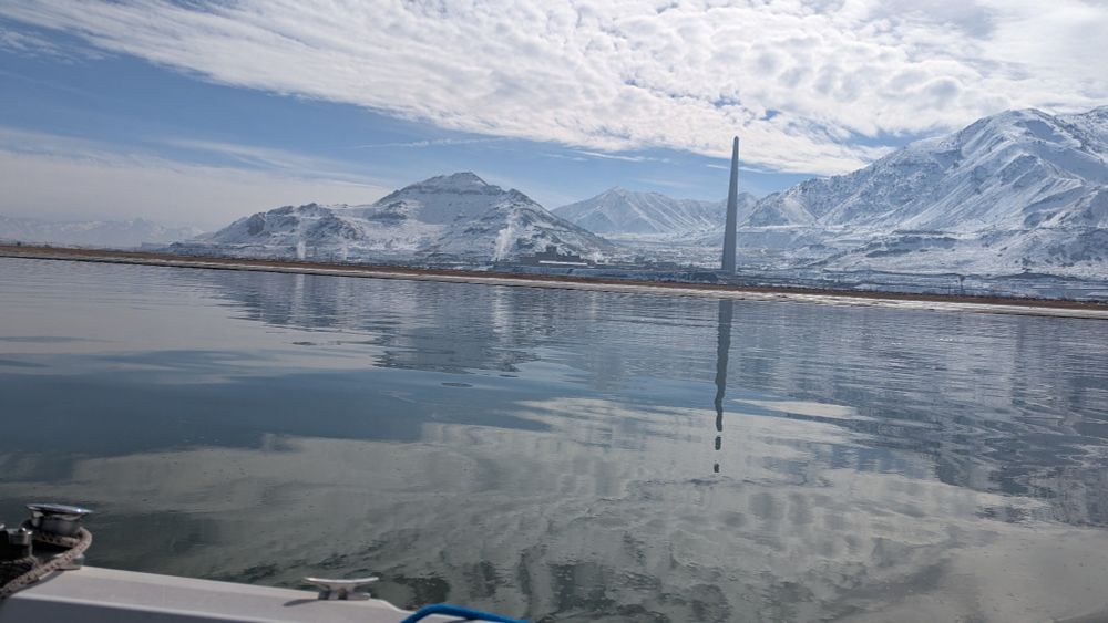 A picture taken from a sailboat. The edge of which can be seen in the foreground. Off the boat the water is glass calm all the way to snow covered mountains. A smokestack rises up from the shore, though no smoke is emitted.