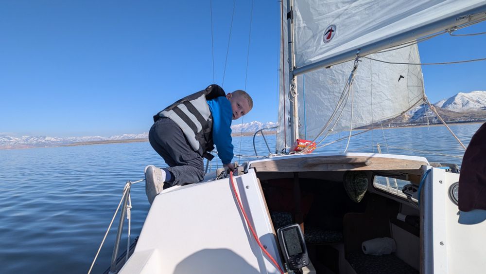 A young boy in a life jacket crawls along the top of the sailboat under sail. Snow capped mountains line the horizon.