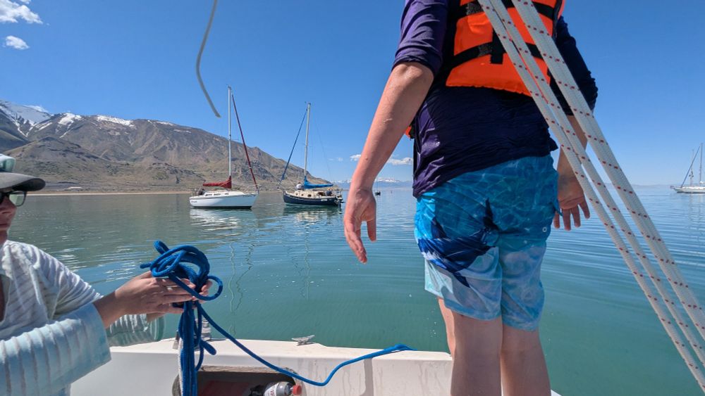 A teenager stands on the seat of a sailboat ready to climb into the water again. Two sailboats float in water nearby.