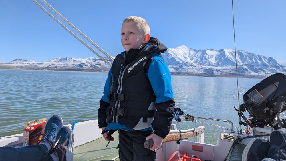 A young boy stands at the tiller of a small sailboat as he watches into the distance. Snowy mountains line the coast.