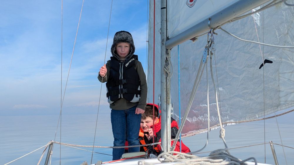 Two children on a sailboat under sail. They are making funny faces. The boat is sailing through still water in the cold with fog in the distance.