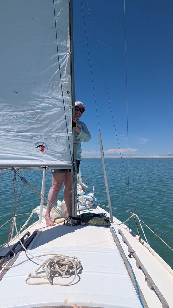 A sailboat approaching a beach under sail. A woman stands in front of the mainsail in shorts and a long sleeve sun shirt. 