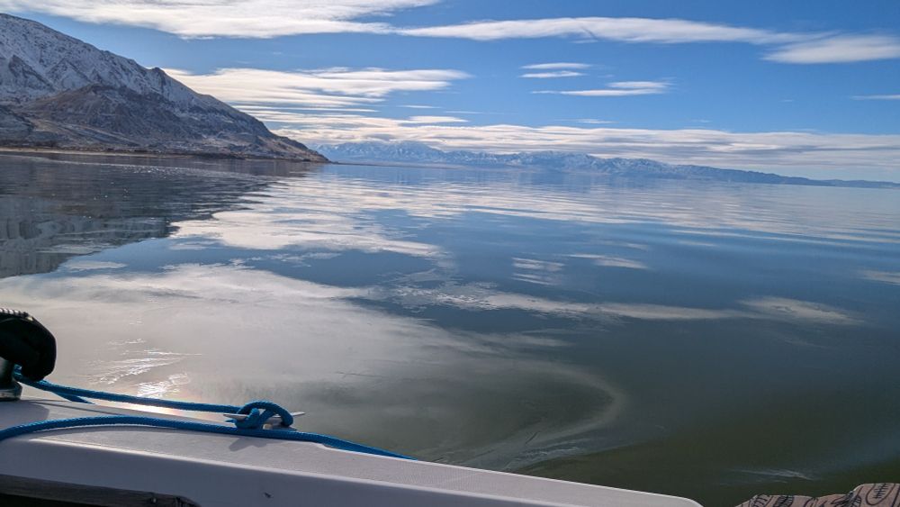 A view from a sailboat. The water is glass calm and a thin layer of ice lays in spots on the water. The sky is bright and reflects the mountain down onto the water.