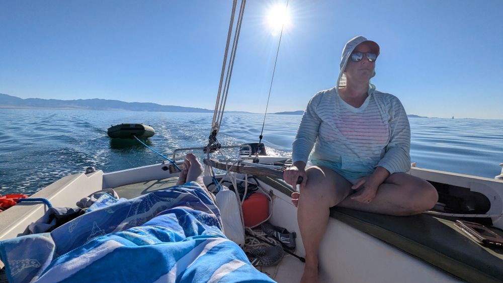 A woman sits at the tiller of a sailboat. The sun is starting its journey to the horizon. A dinghy is at tow behind the boat.