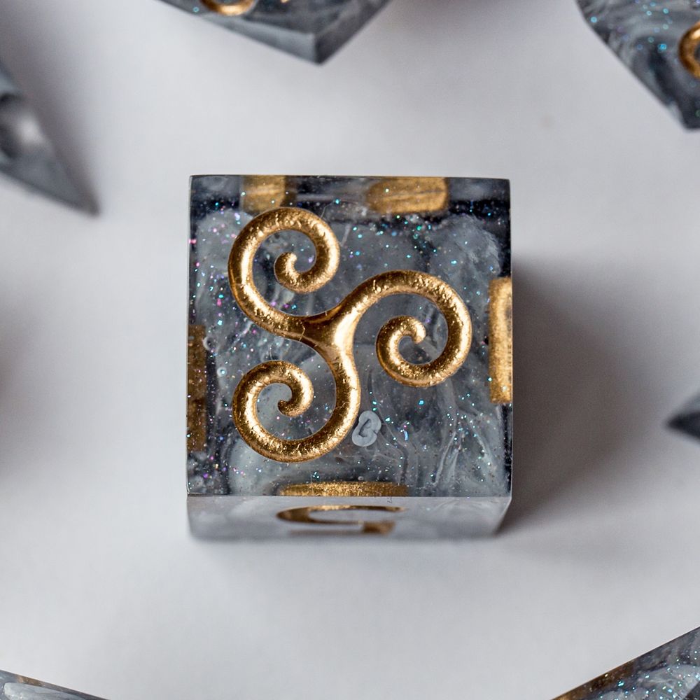 Detailaufnahme eines D6-Würfels auf weißem Hintergrund. Die Seite mit der Zahl 6 zeigt eine goldene Triskele. Der Würfel hat eine graue Betonoptik mit goldenen Ziffern.
