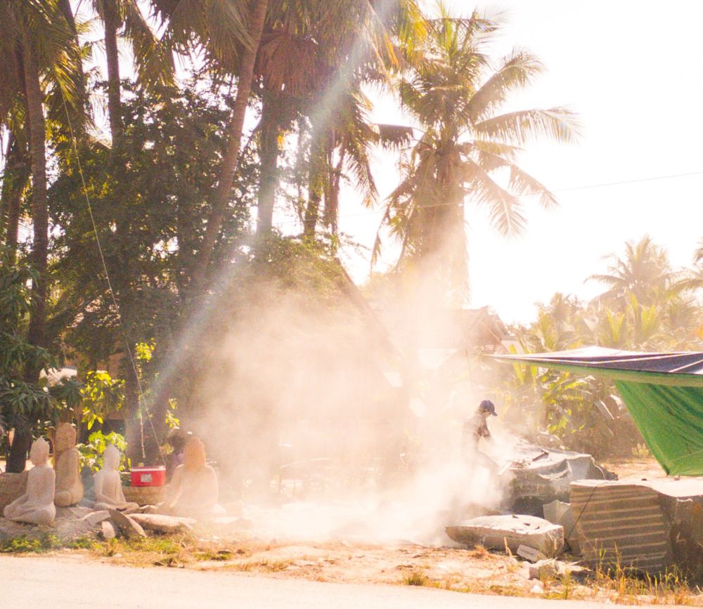 Photo shows a man in a stony dust cloud working on a stone sculpture with a machine tool. It is a sunny day, and the sun plays with the contrasts inside the stone dust. Around the cloud and in the background, there are some sculptures in different modes near finish. To the left there is some large trees, and far in the background there is a low building.  A red fridge box is visible in the shadows under the trees. 