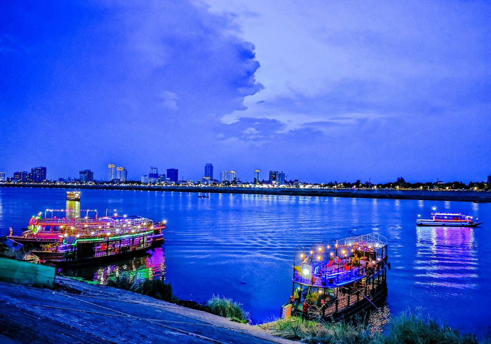 Photo shows two larger double floors restaurant boats on a river with lights and guards ready for an evening cruise on the river. The light is deep blue just after sunset. In the background there is the other river brim, with smaller lower houses and some larger high rises. In the north background the sky is divided in two. To the right it is light blue with few clouds lit up yellow from the sunset. To the left there are dark, stormy/rainy clouds. One of the clouds can seem to be forming an angry man’s profile. There are high expectations for a rainy night.
