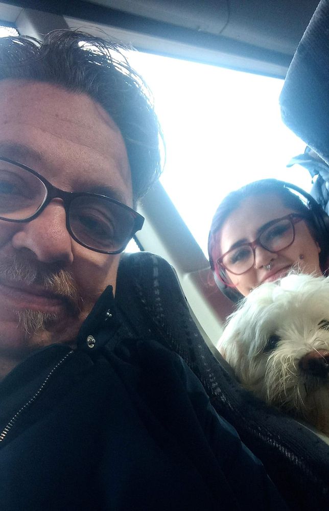 A selfie taken on a bus showing me in glasses and a dark jacket smiling slightly in the foreground, with my daughter in glasses and headphones behind me, smiling while holding a small white fluffy dog. Bright daylight comes through the window behind them.