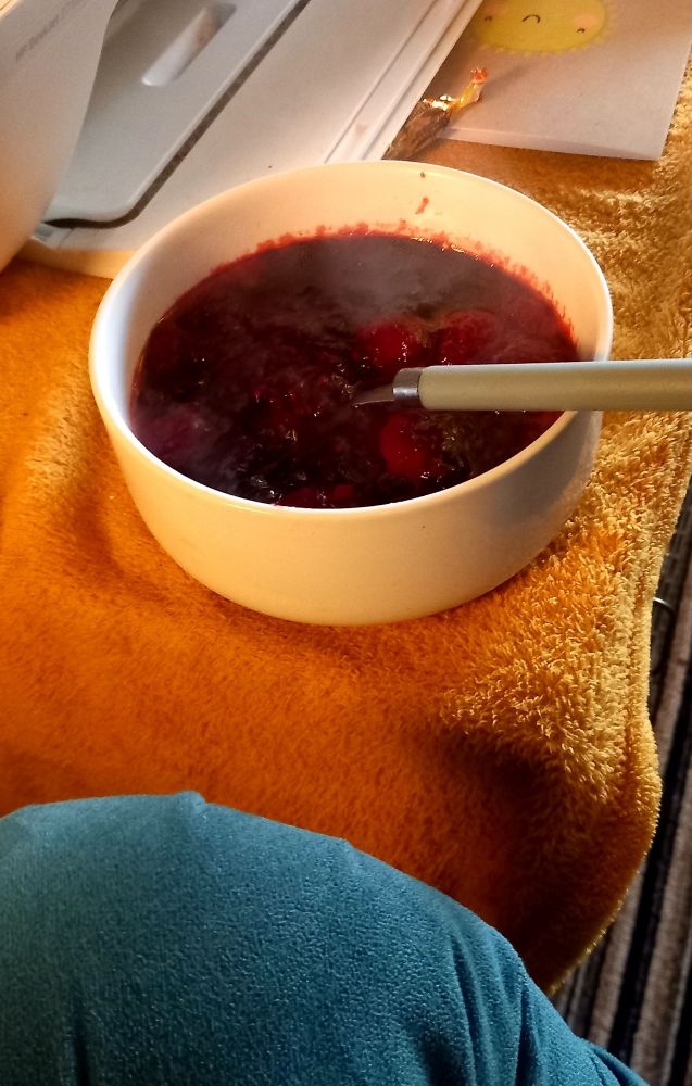 A white bowl filled with a hot, dark red fruit mixture—some berries—sits on a soft orange fabric surface. Steam rises from the bowl, and a spoon rests inside. Nearby are some papers and part of an electronic device, with a small yellow notepad visible in the background.