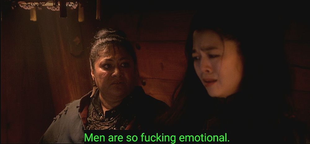 A screenshot from ofmd of Auntie & Zheng stood in a room on a ship. Auntie is looking at Zheng while Zheng looks kind of confused/disgusted at the Revenge crew (off screen). The subtitles 'Men are so fucking emotional.' Is in green at the the bottom