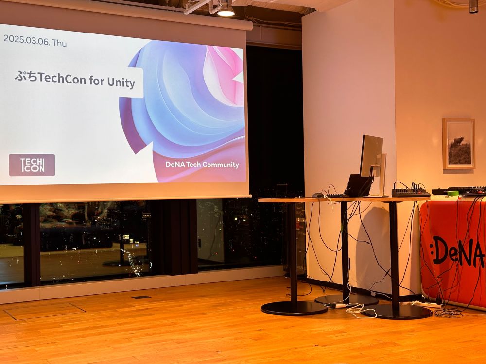 A conference room with a projector screen displaying ‘ぷちTechCon for Unity’ hosted by the DeNA Tech Community. The screen has a sleek blue and pink swirl design. In the foreground, there are two standing desks with laptops, cables, and audio equipment. A city skyline with lights is visible through the large windows in the background