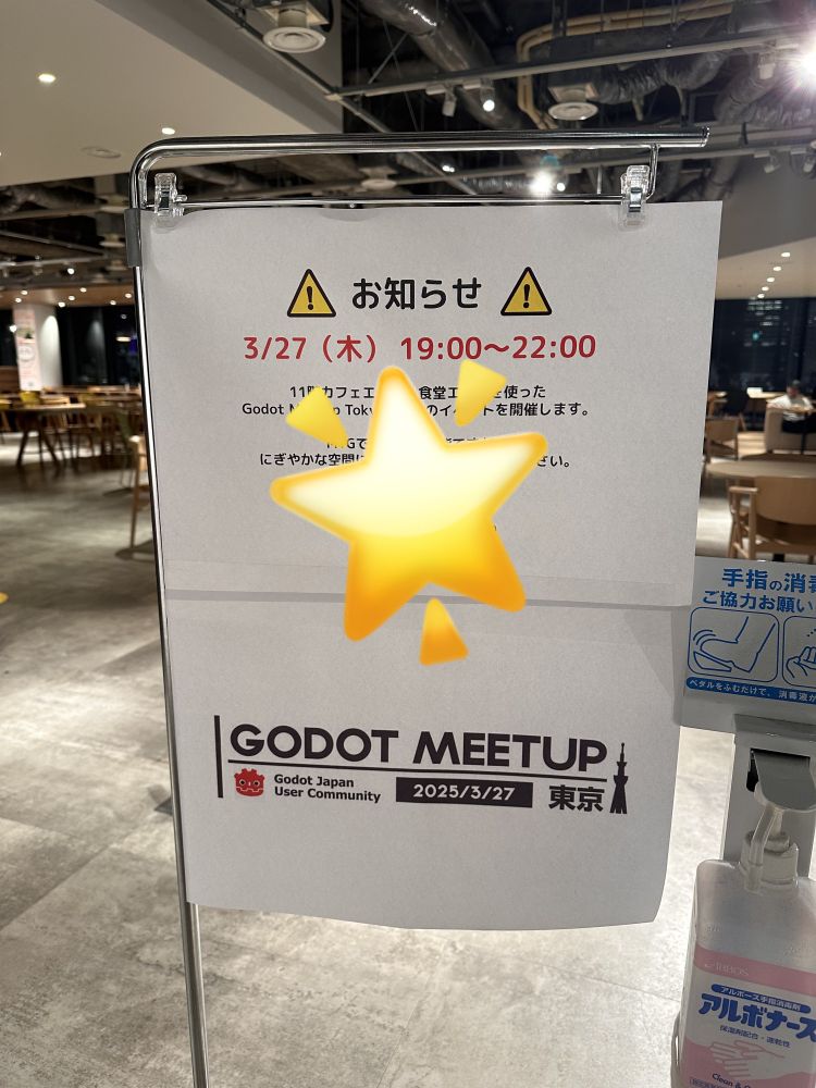 This image shows a printed sign announcing a Godot Meetup event in Tokyo, organized by the Godot Japan User Community. The sign is clipped to a metal stand in an indoor space with modern lighting, wooden tables, and chairs.