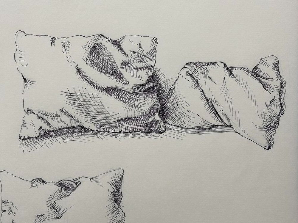 A pen-and-ink drawing of two crumpled pillows on a flat surface. The artist uses hatching and cross-hatching techniques to create shading, depth, and texture. The fabric folds and creases are carefully rendered, giving a sense of softness and volume. A partial sketch of another pillow is visible in the lower part of the image