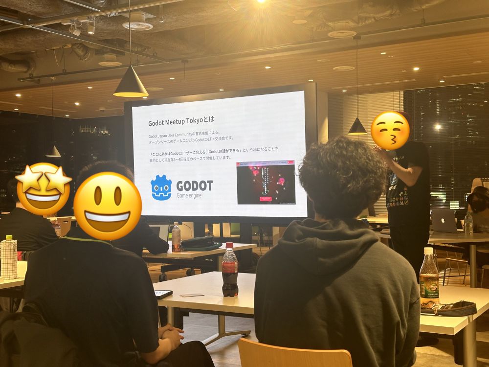 This image shows a Godot Meetup event in Tokyo. A presenter stands in front of a large screen displaying a slide about the event, which describes the Godot Japan User Community and its goal of bringing together Godot users. The slide features the Godot logo and a screenshot of a game. The audience, seated at tables, listens attentively. Some attendees have their laptops open, and drinks are placed on the tables. The setting is a modern, well-lit indoor space with wooden paneling and pendant lights. Several people's faces are covered with emoji stickers, expressing excitement and happiness.