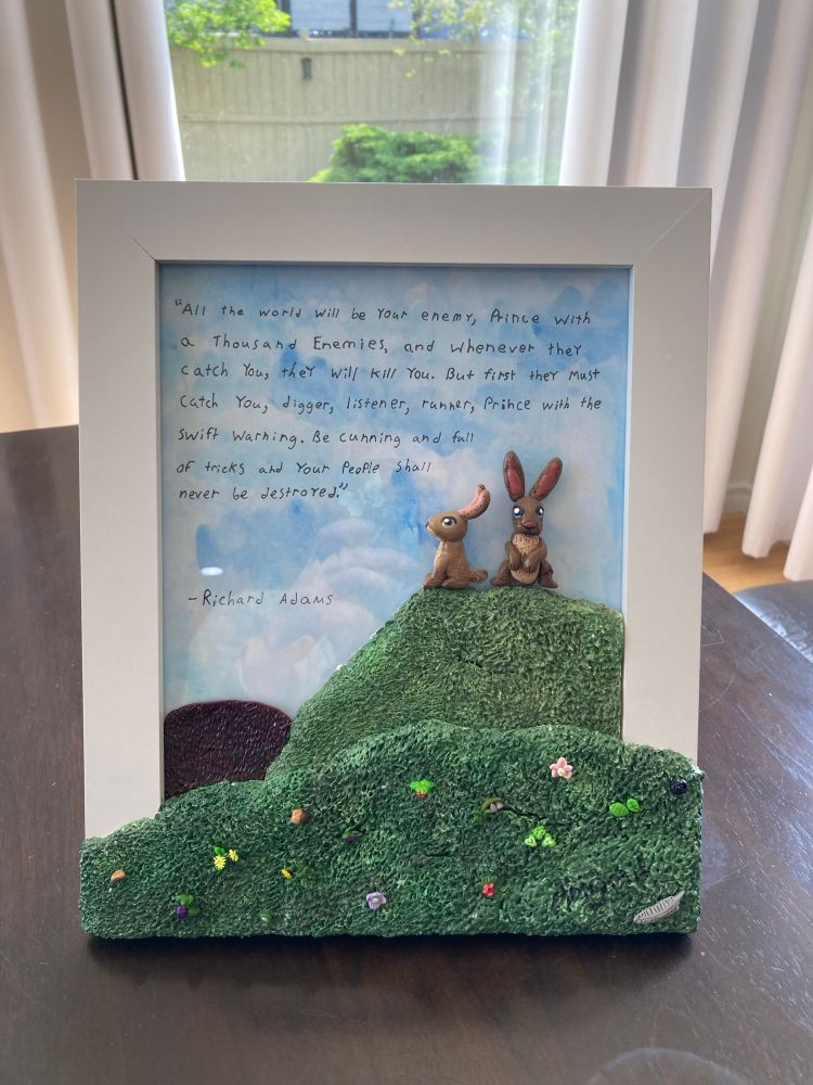 An image of a picture frame with clay made grassy hills spilling over the sides. Two polymer clay rabbits sit atop the hill which is decorated with polymer clay made flowers and plant life. Behind the rabbits is a water coloured blue sky with a quote from the book Watership Dowb written on it with black marker. 