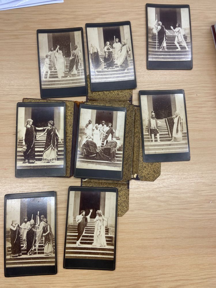 Image of a eight old black and white pictures on a table, showing actors in various positions, gesturing, dressed in Ancient Greek robes, standing in front of a black door opening with a staircase