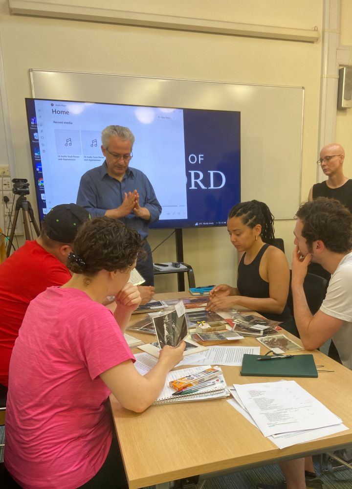 Image of a teaching room with a digital screen at the background, a middle age man in front of it explaining something, and a group of students at a table handling archival material