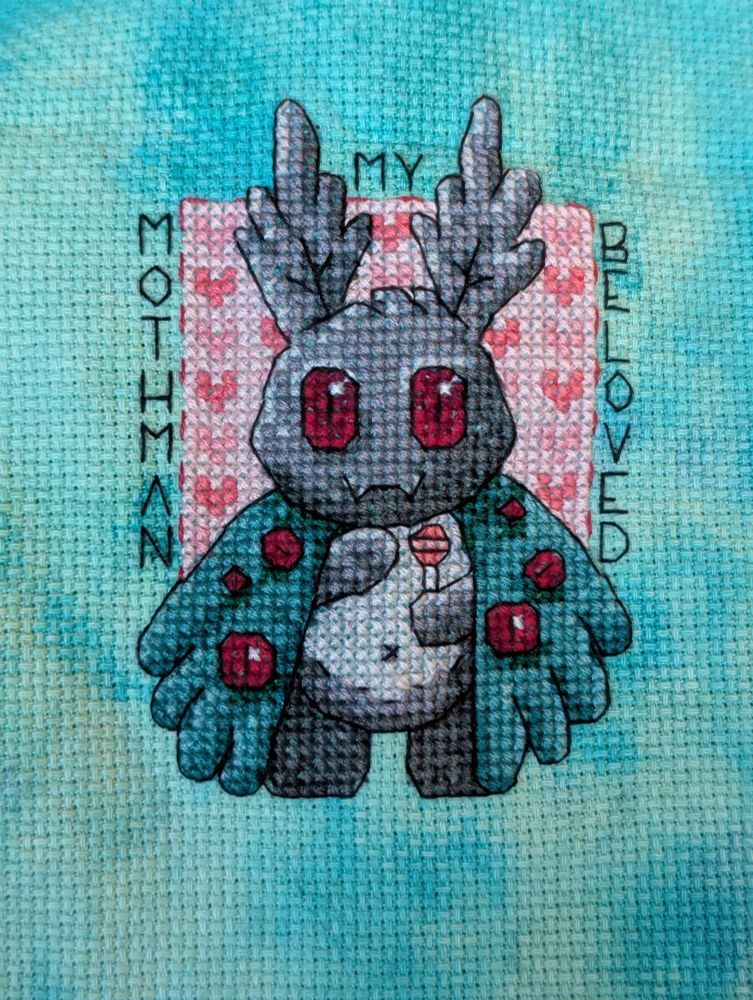 Cross stitch of cute baby Mothman holding a sucker and smiling, with a pink heart patterned background. Text reads Mothman My Beloved 