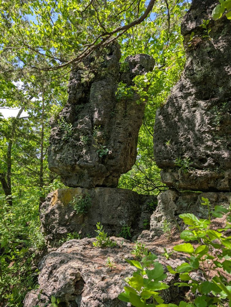 Interesting rock formation, looking like a big boulder atop a smaller boulder on the edge of a cliff. Rocks are surrounded by greenery from tree branches 
