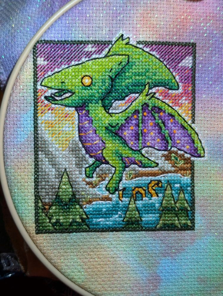 A cross stitched plushie pterosaur with bright green body and a purple belly and wing membrane. It is flying above mountains, evergreens, and a body of water with an orange sea serpent. The sky behind the pterosaur is sunset colors fading from yellow at the horizon to a deep purple at the top, with small pale pink clouds.