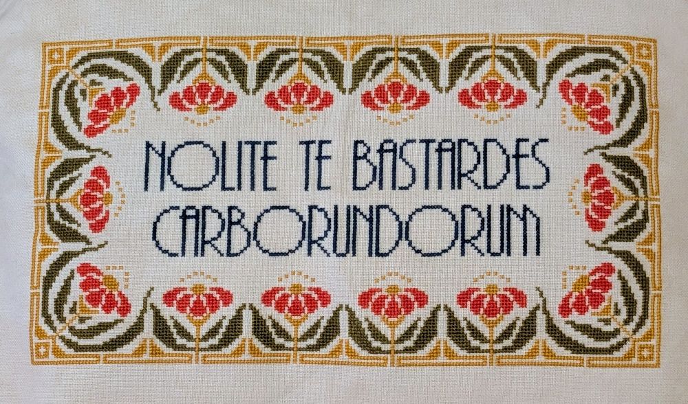 Cross stitch reading "Nolite te bastards carborundorum" in dark blue with a floral border in red-orange, green, and gold